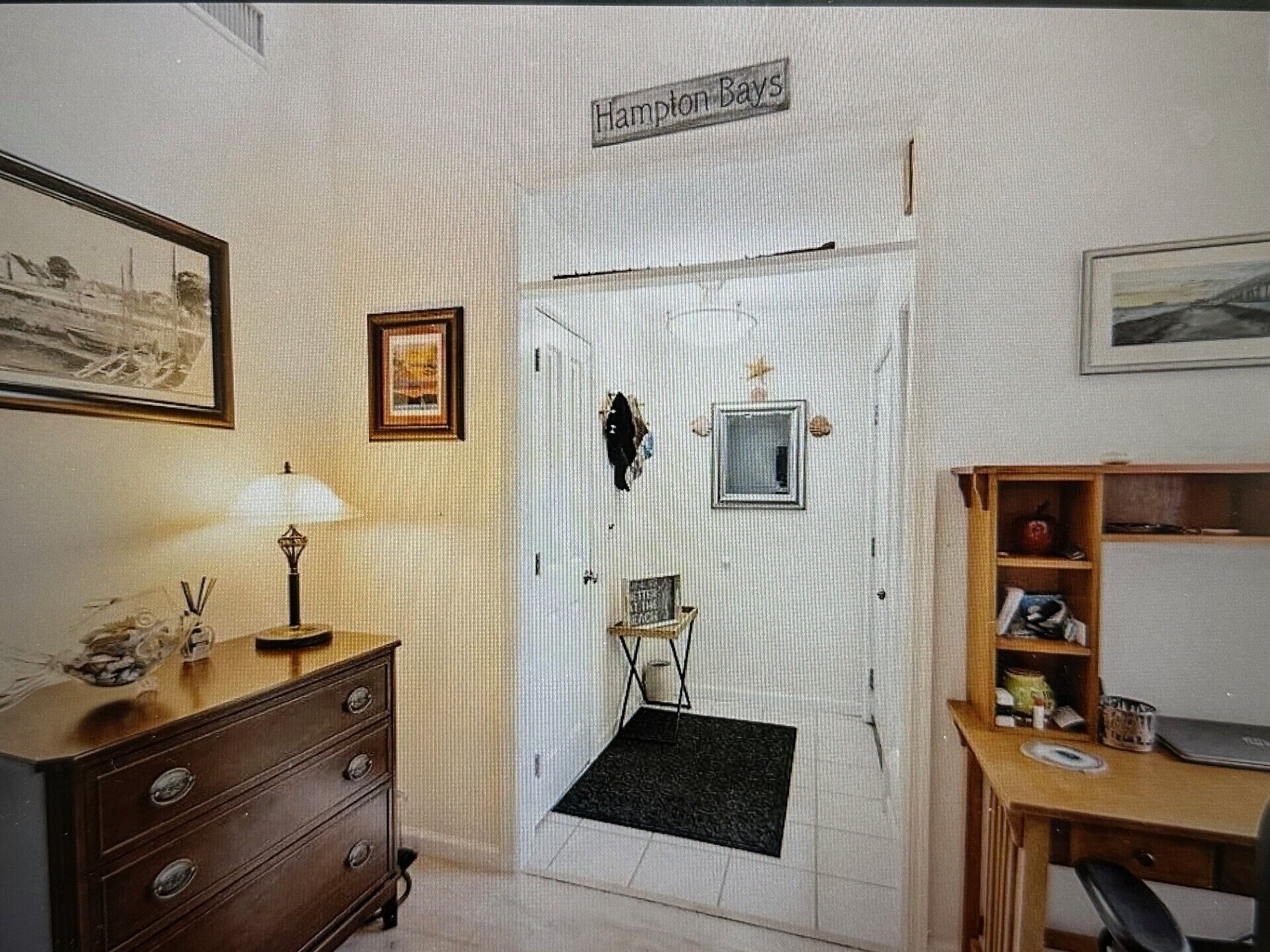 A room with a wooden dresser, a desk, a framed picture on the wall, a small window, and a door labeled 'Hampton Bays.'
