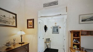 Una habitación con un escritorio de madera, una silla, una lámpara, un aparador, un cuadro enmarcado, una mesita con una silla y una puerta con un letrero encima.