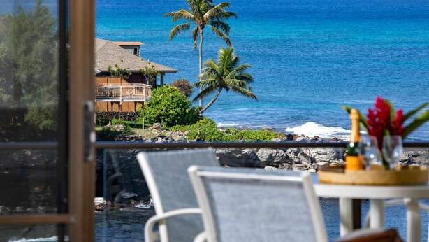 A coastal view with a house, palm trees, and a table with a bottle and flowers. - Napili Point | Oceanfront 3 BR, Sleeps 8 | Car Incl. w/ 6+ Nights | NAP ML-1556 by KBM (Lahaina)