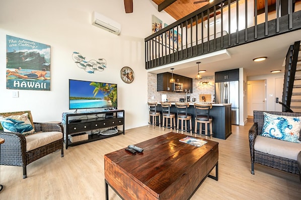 A spacious living area with a TV, wooden coffee table, wicker furniture, a kitchen with bar stools, and a staircase.