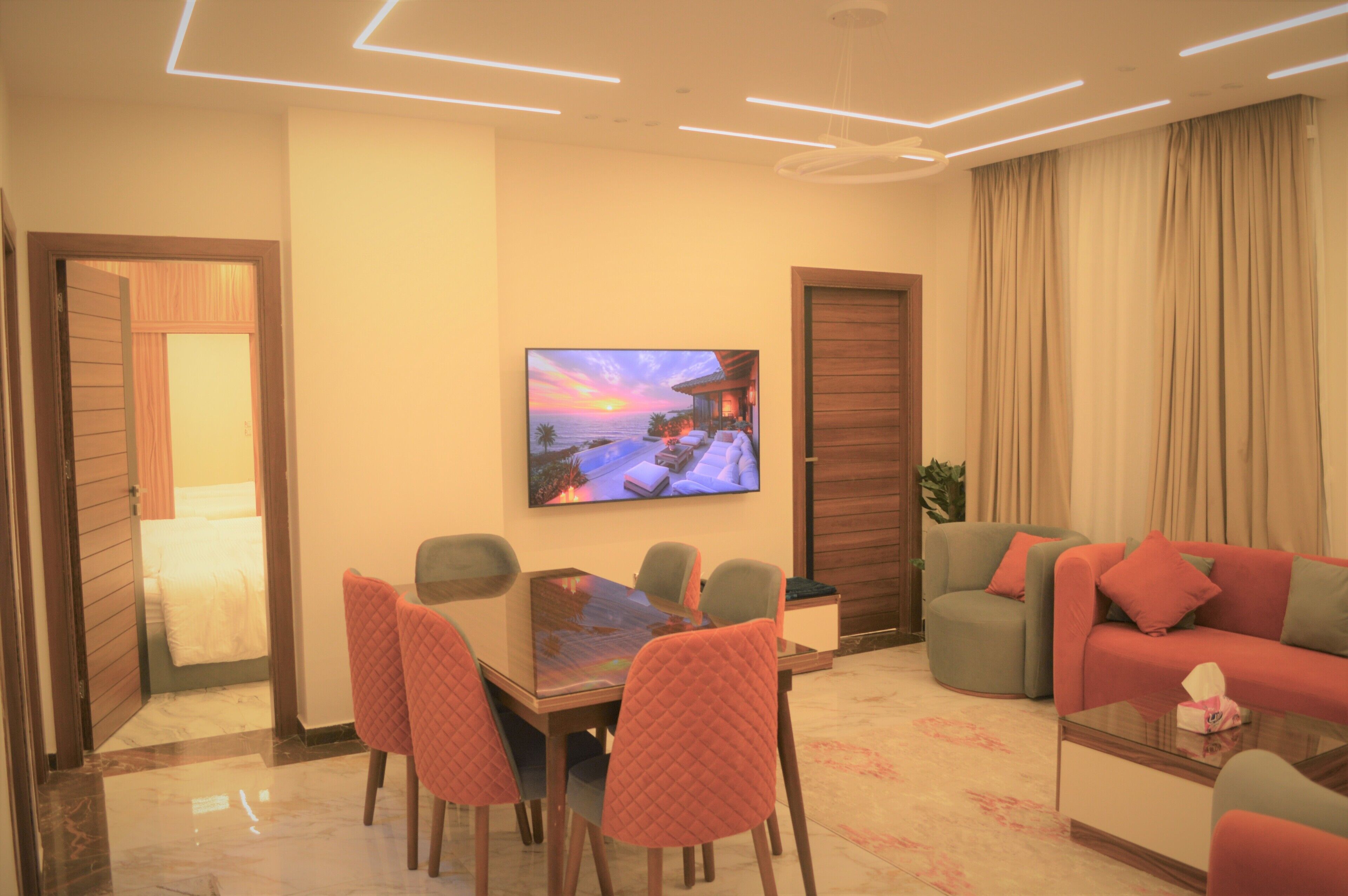 A modern living room with a dining area, a TV mounted on the wall, and a large window with curtains.
