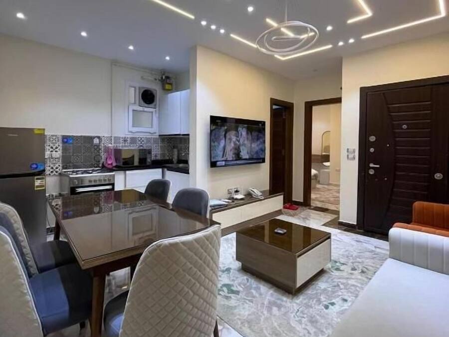 A modern living room with a dining area, a TV mounted on the wall, and a kitchen in the background.