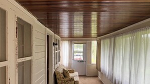 A room with a wooden ceiling, a floral sofa, a bed with a purple blanket, and a window with sheer curtains.