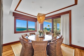 A dining area with a round table, wicker chairs, a hanging light fixture, and large windows with a view of the ocean.
