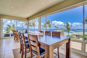A dining area with a large table and chairs, large windows with a view of the ocean and palm trees.