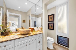 A bathroom with a large mirror, a bowl sink, white cabinets, and a window with blinds.