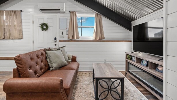 Un acogedor espacio tipo loft con un sofá de cuero marrón, una mesa de centro de madera, un televisor de pantalla plana sobre un soporte de madera y un amplio ventanal con cortinas.