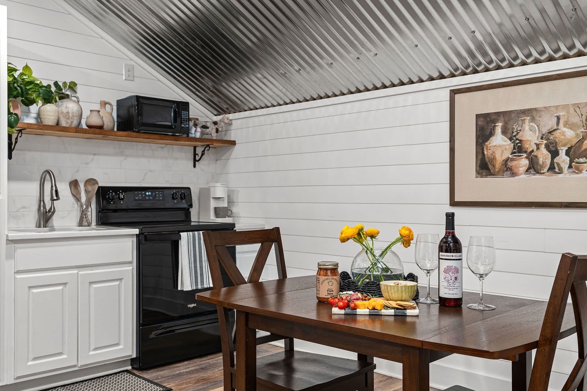 Une cuisine avec des meubles blancs, une cuisinière noire et une table en bois agrémentée de fleurs, d’une bouteille de vin et de verres.
