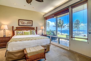A bedroom with a large bed, two bedside tables with lamps, a wooden bench, a large window with a view of palm trees and the ocean, and a framed picture on the wall. - Kapalua Bay Villas | 2 Free Rental Cars | KBM Resorts | Premium Bay Villa | 2 Units, 3 Bedrooms | ML-1604 (Lahaina)