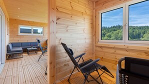 Interior de una cabaña de madera con sofá, sillas, una mesa y un amplio ventanal con vistas al bosque.