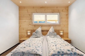 A double bed with a patterned bedspread, wooden walls, a window, and bedside lamps.
