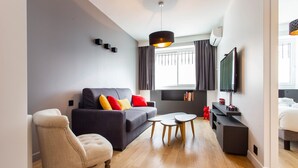 Ein modernes Wohnzimmer mit Sofa, Sessel und Couchtisch, einem an der Wand montierten Flachbildfernseher und einem Fenster mit Vorhängen.