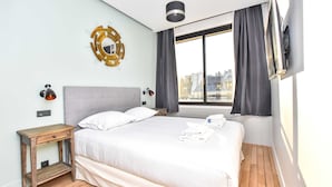 Una habitación de hotel moderna con una cama grande, una mesita de noche, un televisor montado en la pared y vistas a la ciudad a través de la ventana.