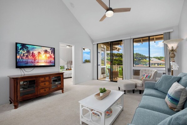 A modern living room with a flat-screen TV, a wooden cabinet, a white coffee table, a blue sofa, and a large window offering a view of the outdoors.