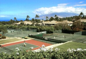 Un complexe de courts de tennis comprenant plusieurs terrains, entouré d’une végétation luxuriante et de palmiers, avec une vue sur une montagne et l’océan en arrière-plan.