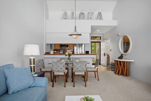 A modern living room with a blue sofa, a dining table with chairs, a kitchen area, and a large 'ALOHA' sign above the kitchen.