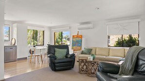 Un salon moderne avec un canapé en cuir noir, un canapé beige et un coin repas comprenant une table et des chaises. Un tableau orne le mur, et une décoration en forme d’étoile y est également présente.
