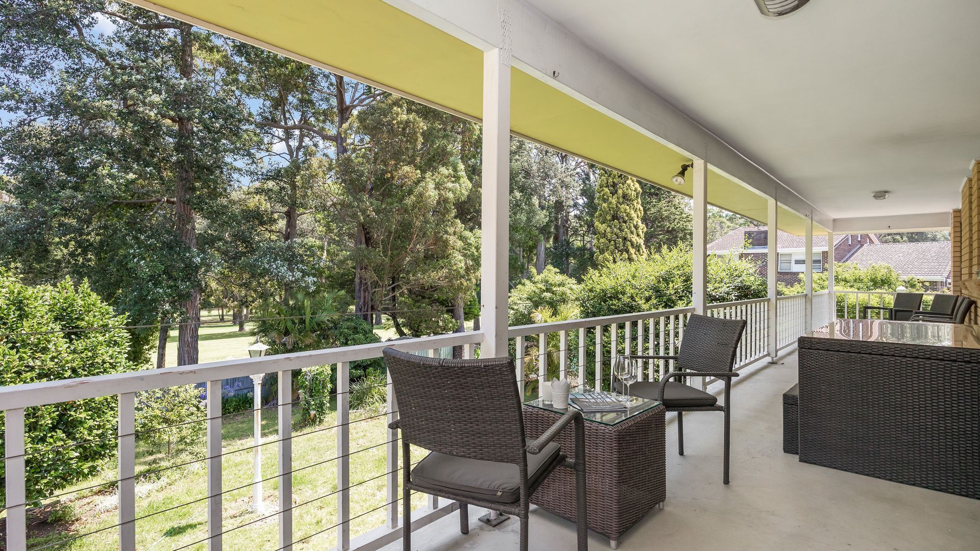 Un balcón techado con mobiliario de mimbre, una mesa de vidrio y vistas a un jardín exuberante.