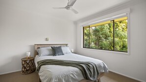 Une chambre à coucher comprenant un lit, une table de chevet, une commode, un ventilateur de plafond et une fenêtre donnant sur des arbres.