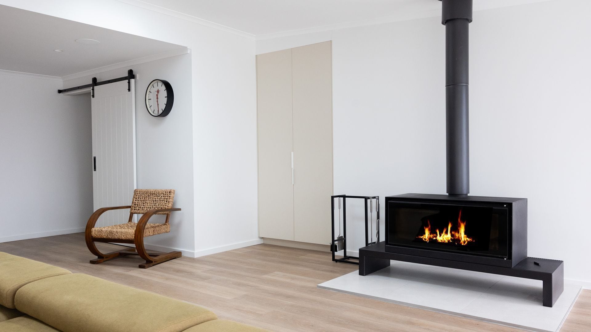 Een moderne woonkamer met een zwart houtkacheltje, een rieten fauteuil en een gele bank.