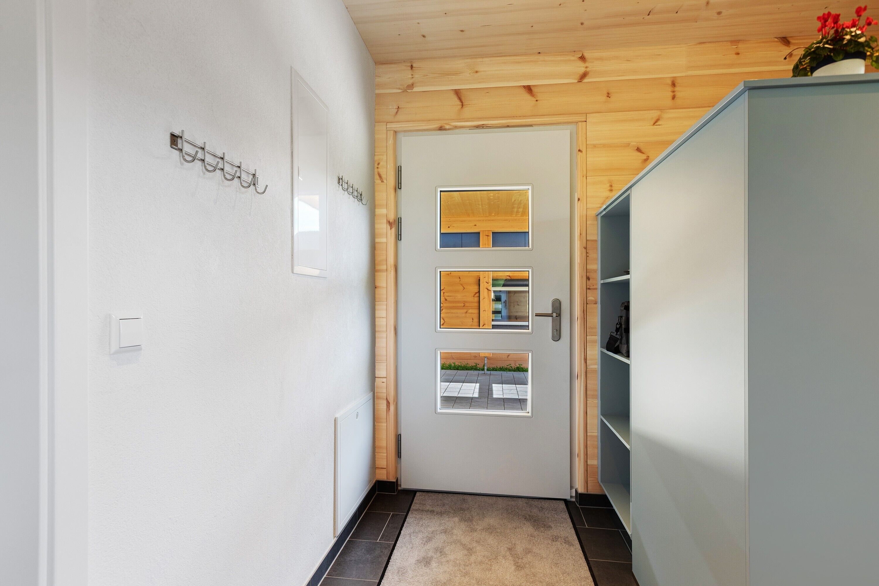 Un corridoio con soffitto in legno, una porta bianca con pannelli di vetro e un mobiletto azzurrognolo con ripiani.