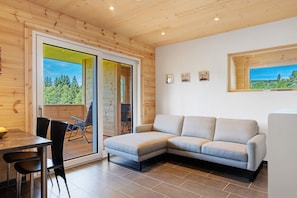 A modern interior with a wooden ceiling, a grey sectional sofa, a dining table with chairs, and a sliding glass door leading to an outdoor deck. - Mettmapark – Chalet 1 with Private Whirlpool & Sauna, Breakfast Included (Grafenhausen)