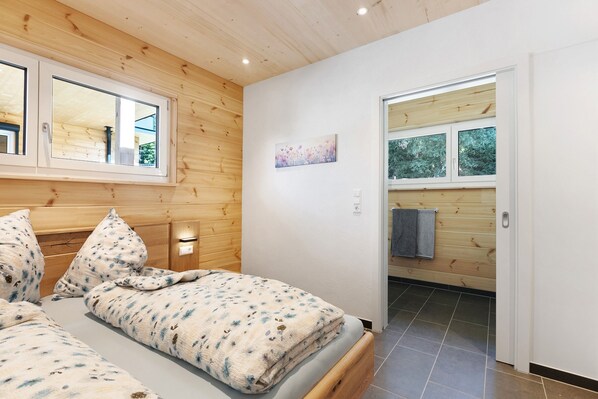 A bedroom with a wooden ceiling, a bed with a floral duvet, a nightstand, and a window.