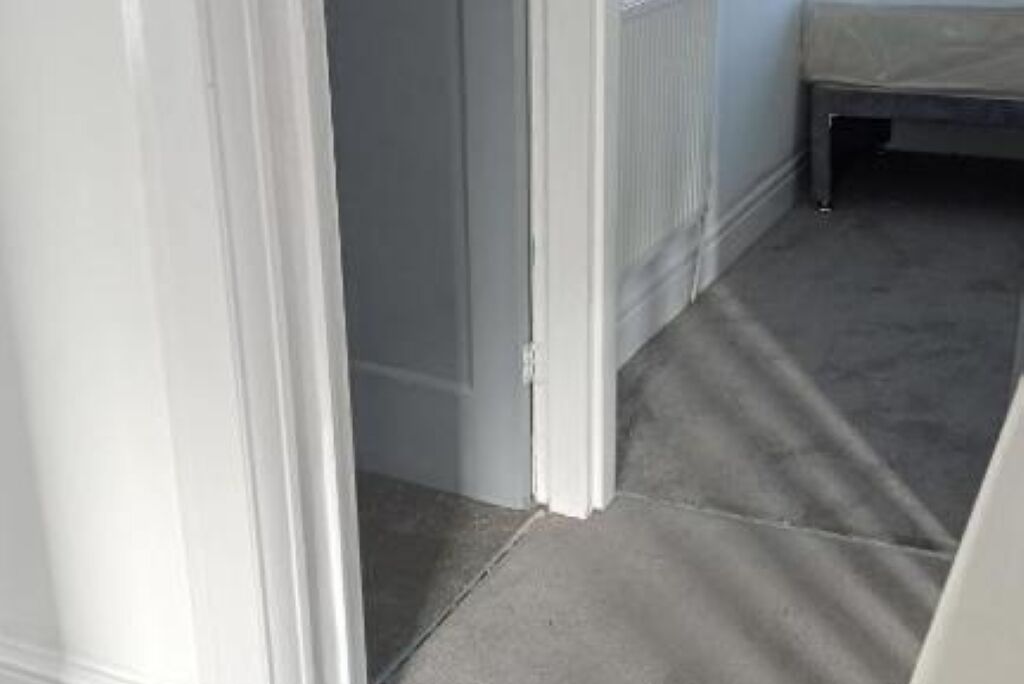 A narrow hallway with a carpeted floor, a radiator, and a bed in the background.