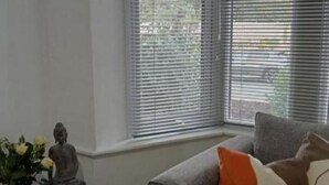 Une pièce avec un canapé gris, une statue posée sur une petite table et une fenêtre avec des stores.