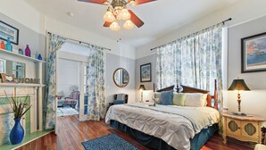A bedroom with a wooden floor, a bed with a white and blue bedspread, a nightstand with a lamp, and a ceiling fan. - Historic Charm in Grand Gulfport Home; walk to beach, casino, port, aquarium (Gulfport)