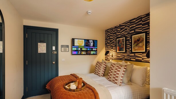 Une chambre à coucher avec un lit, un téléviseur fixé au mur, une porte et un mur décoré d’un motif.