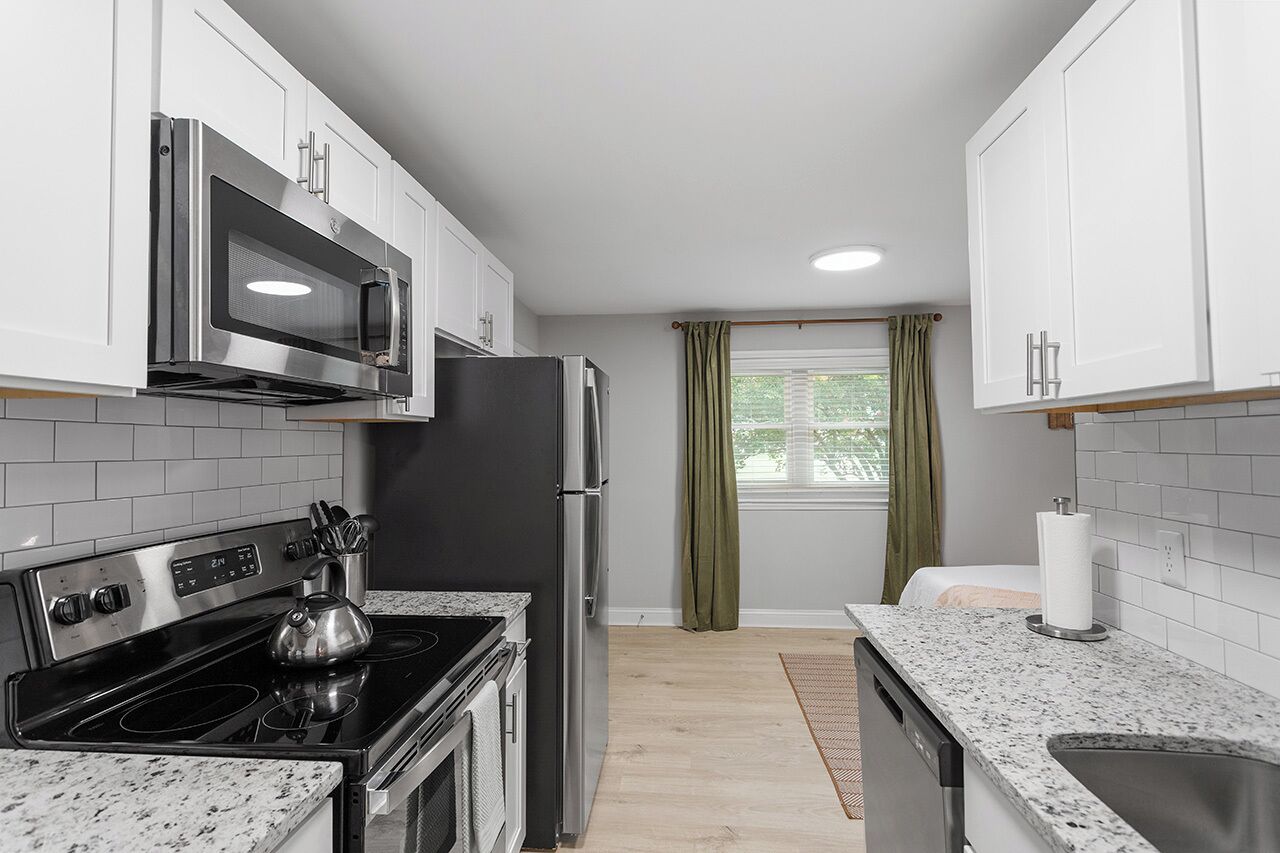 A modern kitchen with white cabinets, stainless steel appliances, a granite countertop, and a window with green curtains.