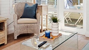 A cozy corner with a wicker chair, a glass coffee table, and a view of greenery through large windows. - Ocean Views At Cottesloe Beach (Cottesloe)