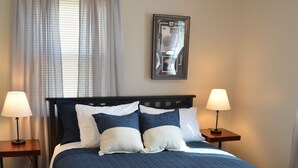 Ein ordentlich bezogenes Bett mit einer dunkelblauen Decke, zwei Nachttische mit Lampen, ein gerahmtes Bild an der Wand und ein Fenster mit Vorhängen.