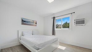 A bedroom with a bed, a window with curtains, an air conditioning unit, and a painting on the wall.