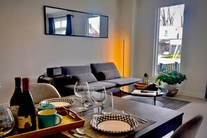 A dining area with a table set for two, a sofa, a potted plant, and a window with a view.