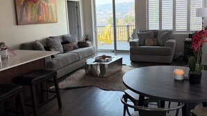 A modern living room with a gray sofa, a coffee table, and a dining area with a round table and chairs. There is a painting on the wall, a ceiling fan, and a view of the outdoors through large windows.
