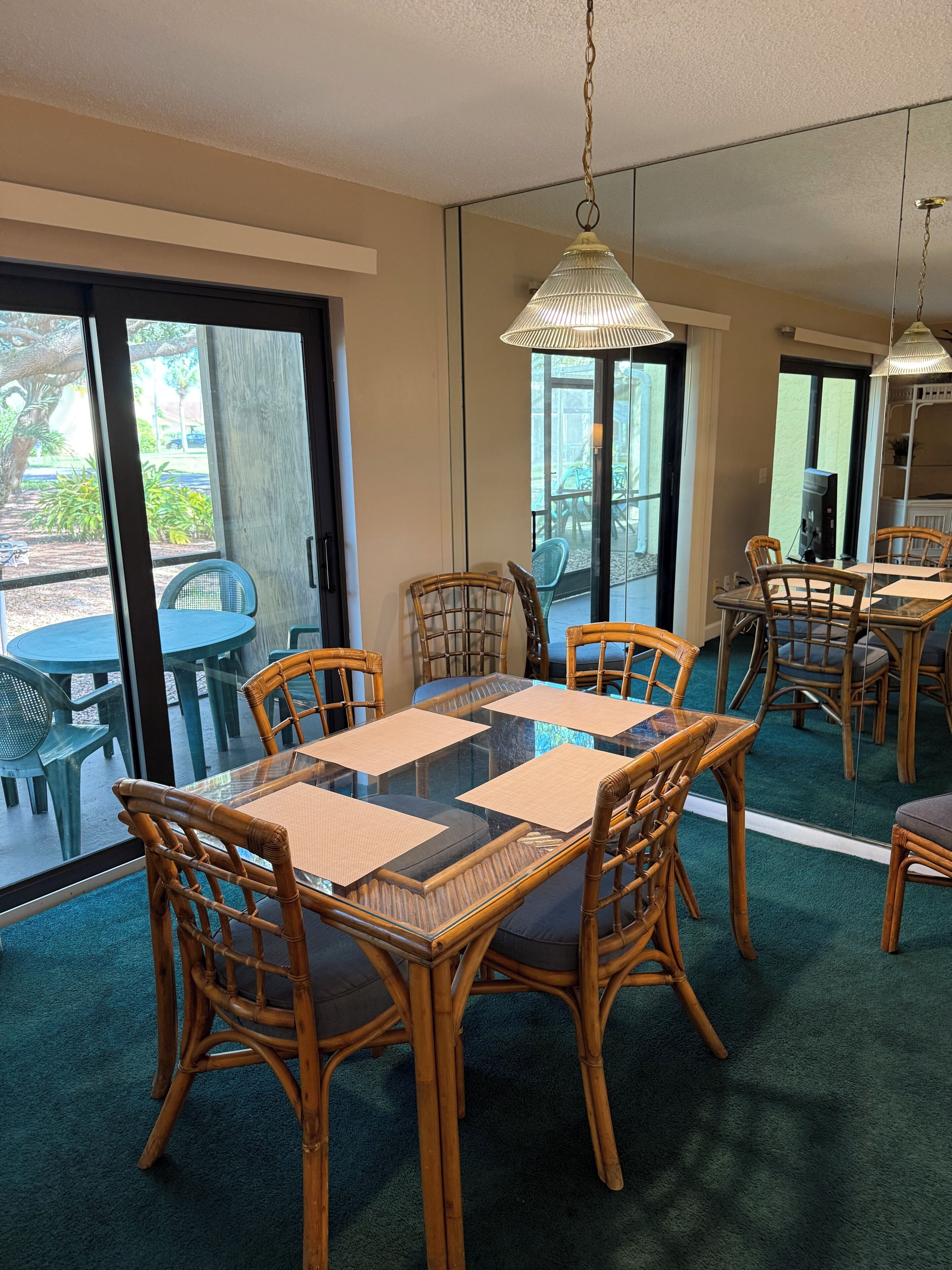 A dining area with a glass table surrounded by wicker chairs, a hanging light fixture, and a view of the outdoors through large glass doors.