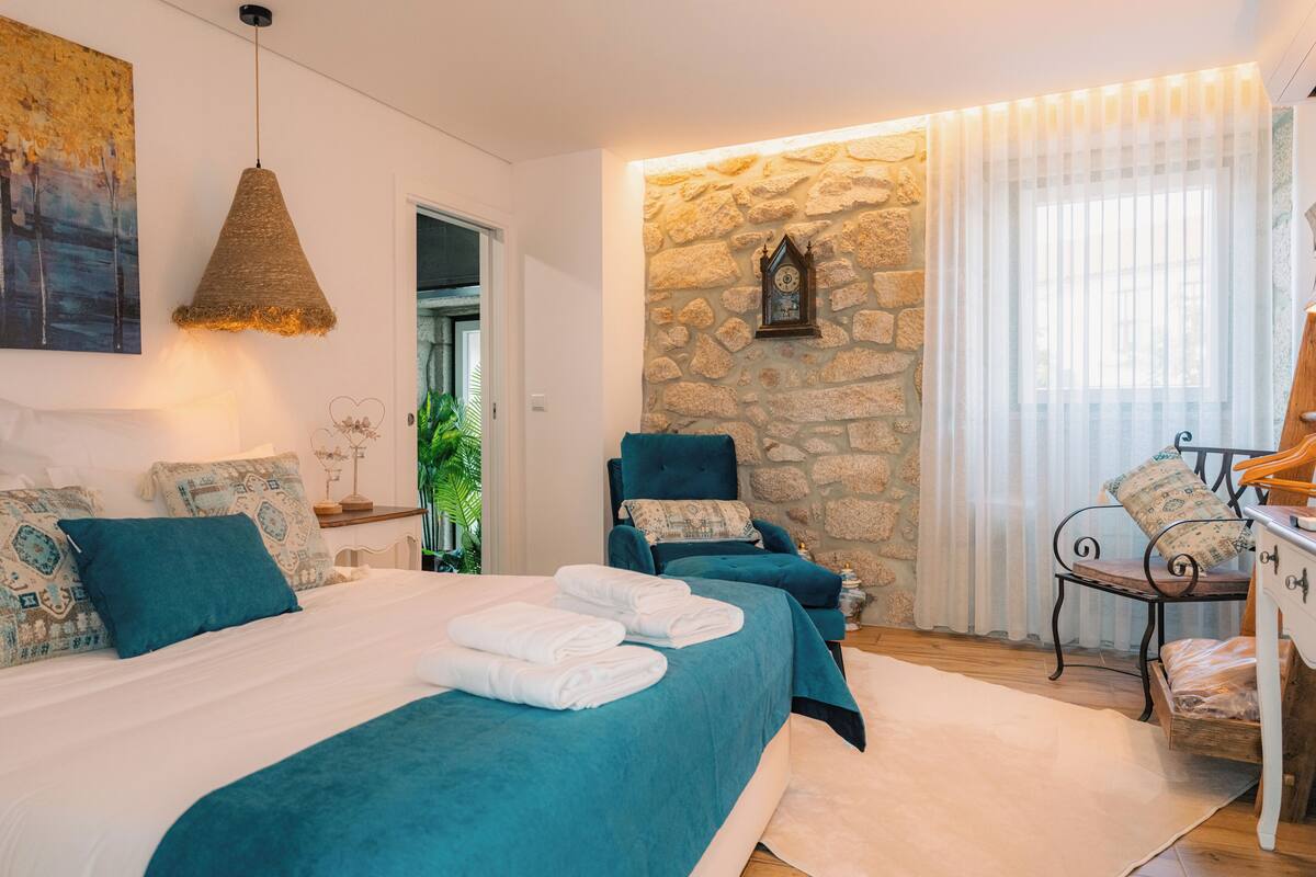 A bedroom with a stone wall, a bed with teal bedding, a chair, and a bedside table.