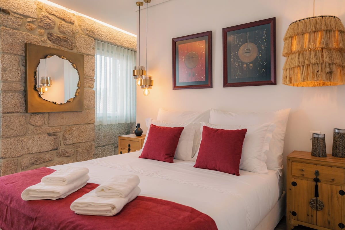A neatly made bed with white and red bedding, a stone wall, a mirror, and two framed artworks.
