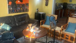 A living room with a black leather sofa, a wooden coffee table, wicker chairs, a painting on the wall, and a kitchen area in the background. - Capricorn by Quest (Cape Maclear)