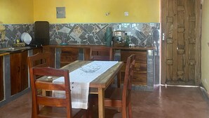 A dining area with a wooden table and chairs, a kitchen with a stone backsplash, and a yellow wall. - Capricorn by Quest (Cape Maclear)