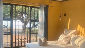 A bedroom with a bed, a window with a view of trees, and a door. - Capricorn by Quest (Cape Maclear)