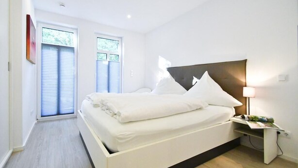 A modern bedroom with a bed, bedside table, and a window with blinds. - "Relaxed vacation days await you in the apartment Sergio 1 on Norderney!" (Norderney)