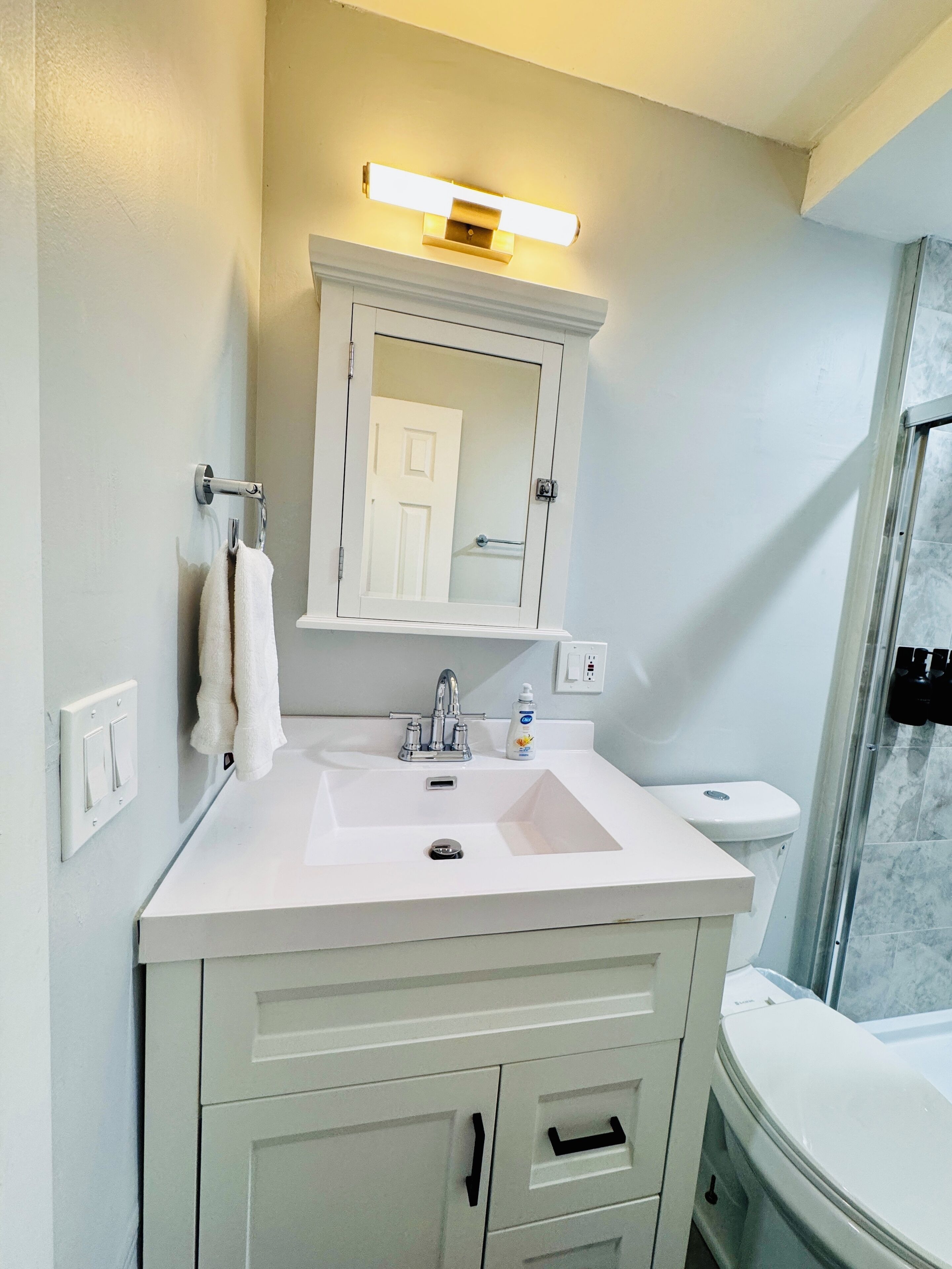 Ein Badezimmer mit einem weißen Waschbecken, einem Spiegel darüber und einer Toilette. Ein Handtuch hängt an der Wand und auf dem Waschbecken steht eine Seife.