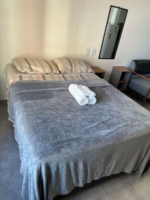 A neatly made bed with a grey blanket and two pillows, a folded towel on the bed, a small table with a lamp, and a chair with a cushion. - Tha flat 311 (Campinas)