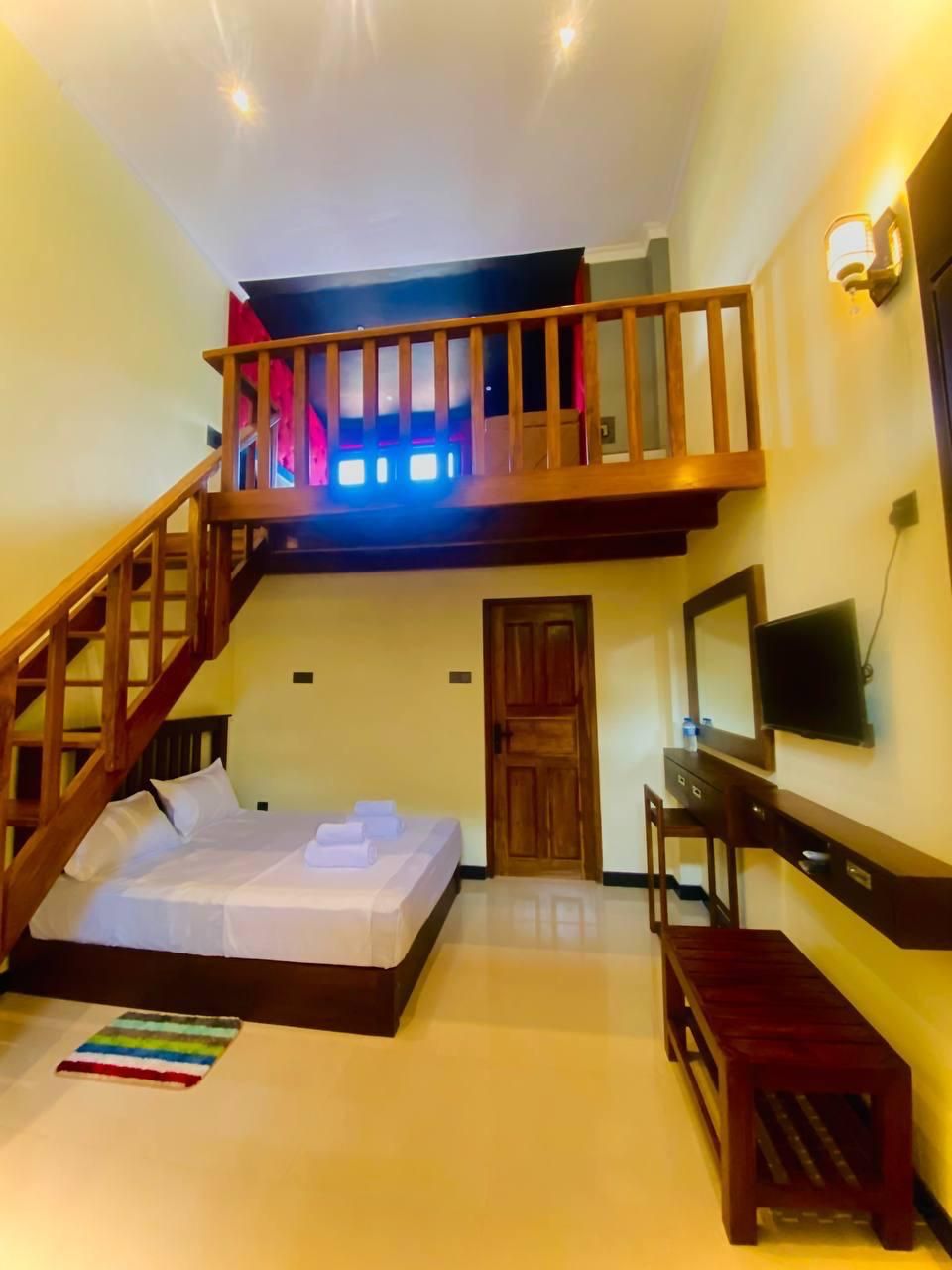 A hotel room with a bed, a desk, a chair, a TV, a wooden staircase, and a balcony with lights.