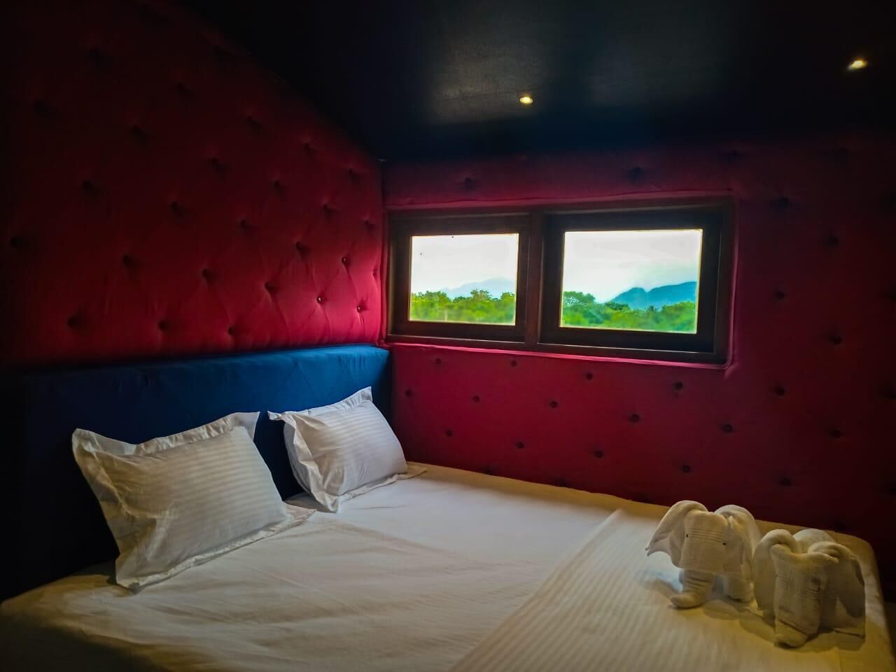 A room with a red and blue upholstered headboard, a large bed with white bedding, and a window with a view of greenery.