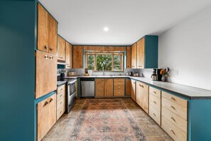 A kitchen with wooden cabinets, a large window, and a patterned rug. - Nucla Haus | In Between Telluride + Moab Basecamp (Nucla)