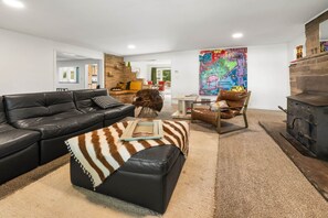A living room with a black leather sofa, a brown and white striped ottoman, a wooden coffee table, a colorful abstract painting, a brown leather armchair, and a stone fireplace. - Nucla Haus | In Between Telluride + Moab Basecamp (Nucla)